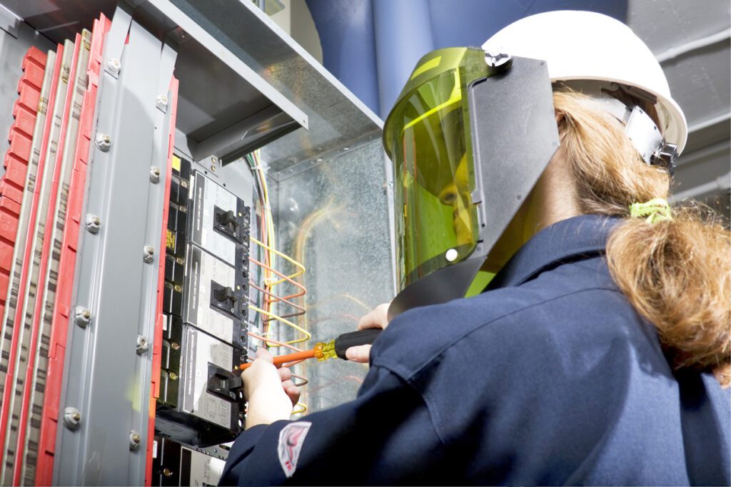 A woman in arc flash clothing, wearing a hard hat and safety glasses, is diligently working on a panel.